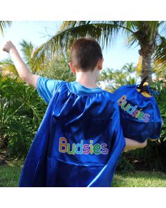 Child wearing bright blue shiny superhero cape with colorful Budsies logo, raising arm outdoors beside palm trees in daytime.